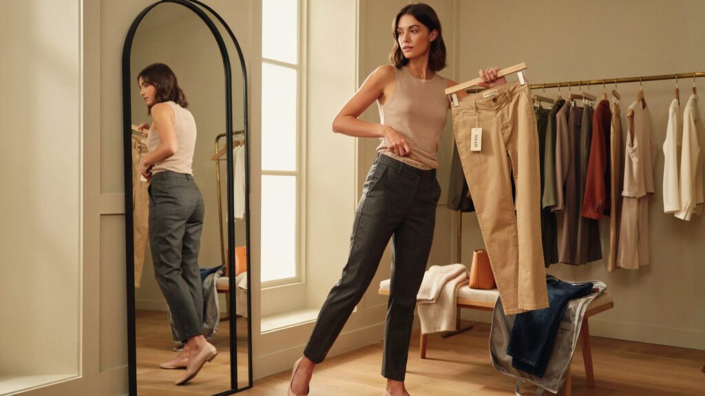 Woman comparing trousers in a fitting room to understand why pants fit differently between brands and how brand-specific sizing affects comfort and fit.